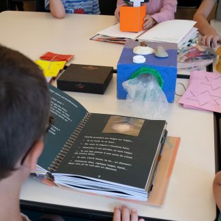 sur une table : livre tactile ouvert devant un enfant, boite avec des vrais fossiles, lunettes de simulation, boite mystère avec éléments à toucher à l'intérieur