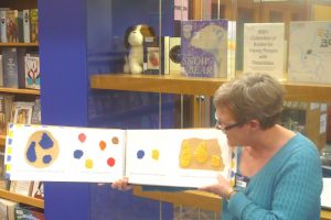 Une femme présente le livre tactile petit bleu petit jaune dans une bibliothèque. C'est un grand livre format paysage avec une reliure spirale.