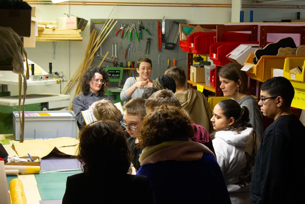 Un groupe d'enfants de dos dans un atelier de bricolage. On aperçoit des outils dans le fond. Deux femmes se tiennent devant le groupe. L'une montre un livre et l'autre fait des signes avec ses mains.