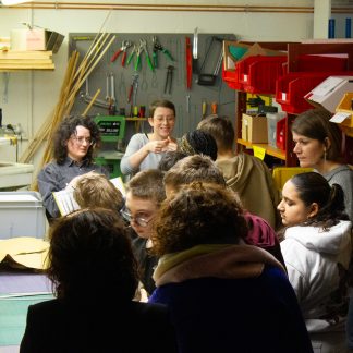 Un groupe d'enfants de dos dans un atelier de bricolage. On aperçoit des outils dans le fond. Deux femmes se tiennent devant le groupe. L'une montre un livre et l'autre fait des signes avec ses mains.