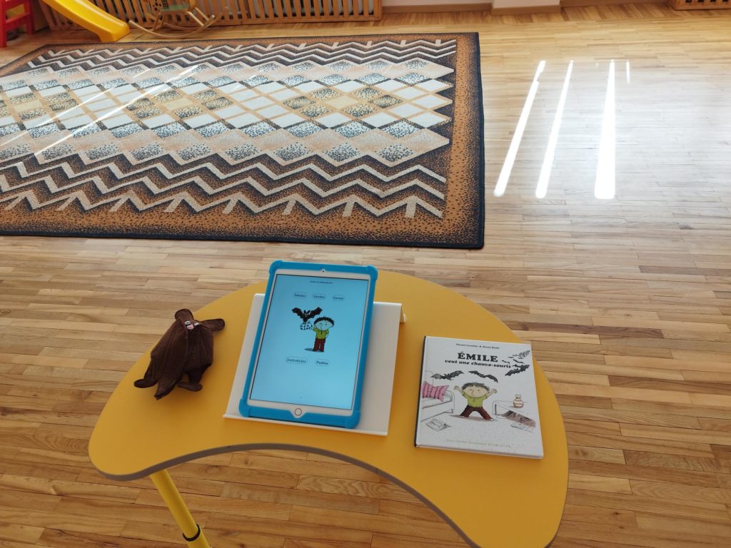 photo qui présente un livre numérique sur tablette avec à sa gauche une petite marionnette de chauve souris et à droite le livre papier dont est issu le livre numérique