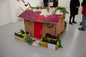 Photo de la maquette du cabanon de l'histoire.Des personnes se tiennent derrière et on peut voir que la cabane est grande (elle arrive au niveau du bassin des personnes debout à côté). Des tuiles en carton ondulé recouvrent le toit. Devant le cabanon sont posées des cagettes avec des faux légumes fabriqués dans des matières variées.