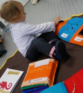 Petite fille qui touche un livre tactile en tissu posé au sol. On voit des clochettes dans le livre