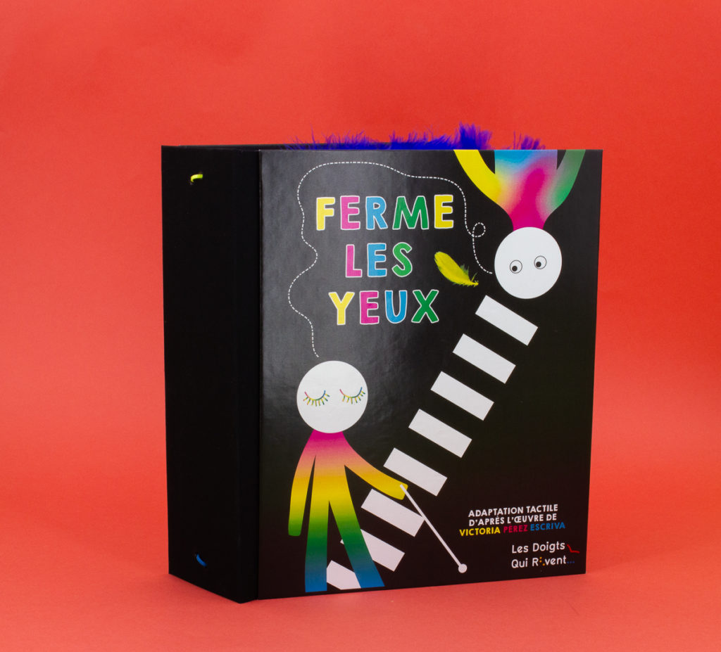 Livre tactile "Ferme les yeux". Il est très épais et de couleur noir. Sur la couverture il y a deux bonhommes multicolores dont un avec une canne blanche