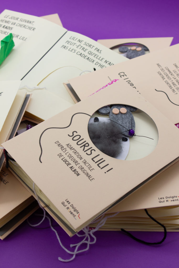 Photo de plusieurs livres Souris Lili empilés. Sur le premier livre, la couverture est percé avec un gros rond à travers lequel on voir le museau d'une souris en laine et nylon.