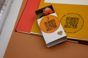 zoom sur une petite boîte avec un QR code dessus et le numéro 3. Il y a une fleur en tissu dans la boîte entrouverte.