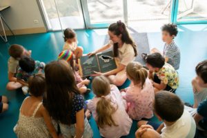 Photo d'une femme assise au sol avec un livre ouvert et une dizaine d'enfants assis tout autour. Sur le livre il y a une illustration tactile d'oiseau.