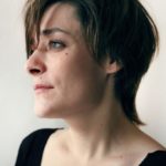 Photo Estelle Faye : jeune femme brune au cheveux courts photographiée de profil. Elle regarde devant elle. Enne ne sourit pas. Son expression est sérieuse et douce à la fois
