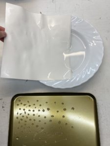 Photo d'une feuille blanche trempée dans une assiette creuse remplie d'eau. On aperçoit à côté un couvercle en métal avec des gouttes de colle dessus.