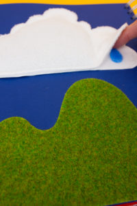 photo d'une illustration tactile du livre avec un gros nuage blanc au dessus d'une colline verte. Un doigts humain soulève une partie du nuage pour trouver en dessous une goutte d'eau