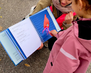 photo d'un livre ouvert avec à gauche du texte noir et braille coloré en bleu et à droite une illustration tactile de calamar. Le livre est tenu par une jeune femme et une petite fille est en train de toucher l'illustration.