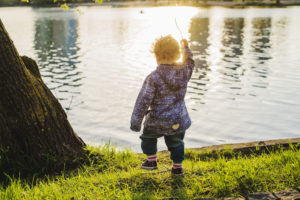 De dos, un petit enfant se tient debout sur l'herbe, devant une étendue d'eau. Il porte des veêtements de pluie et brandit une brindille.