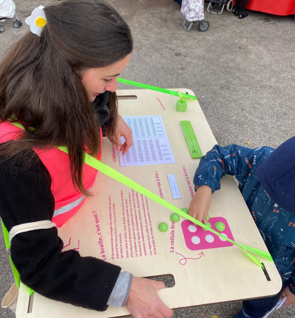 Camille notre animatrice qui déambule dans les rues avec le brailleur ambulant et un enfant essaye d'écrire en braille sur le plateau qu'elle porte